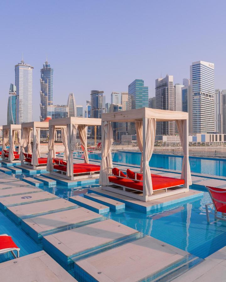 Canal Central Hotel Business Bay Dubaj Zewnętrze zdjęcie The photo features a luxurious setting by a poolside with several white cabana-style structures that have red cushions. Each cabana is draped with sheer fabric, providing a stylish and comfortable lounging area. The background showcases a modern skyl