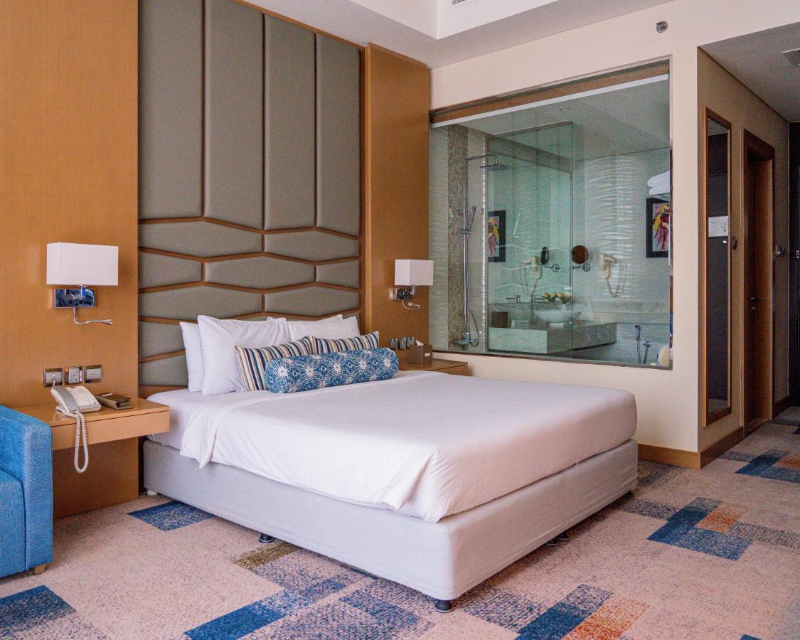 Canal Central Hotel Business Bay Dubaj Zewnętrze zdjęcie The photo shows a modern hotel room featuring a neatly made bed with white linens and decorative pillows. The headboard has a geometric design, and there are bedside lamps on either side of the bed. Next to the bed, there's a small nightstand that in