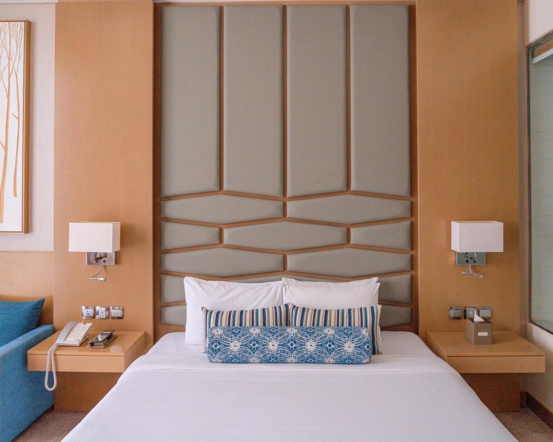 Canal Central Hotel Business Bay Dubaj Zewnętrze zdjęcie The photo shows a modern hotel room with a neatly made bed featuring white linens and a decorative blue pillow. The headboard of the bed has a geometric design and is complemented by soft lighting from wall-mounted lamps on either side. There are nig