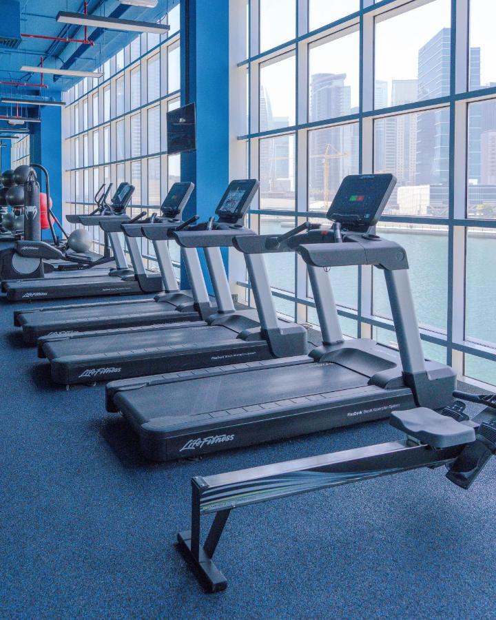 Canal Central Hotel Business Bay Dubaj Zewnętrze zdjęcie The photo shows a series of treadmill machines lined up in a gym or fitness center. The treadmills have digital displays on the front and are situated near large windows that provide a view of the outside. The flooring is carpeted, and the environmen