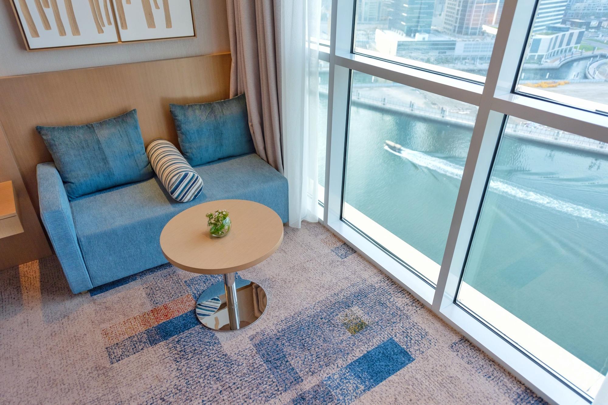 Canal Central Hotel Business Bay Dubaj Zewnętrze zdjęcie The photo shows a modern interior space featuring a cozy seating area. There is a blue sofa with two cushions and a decorative striped pillow. A small round table stands in front of the sofa, topped with a small plant. The large windows provide a vie