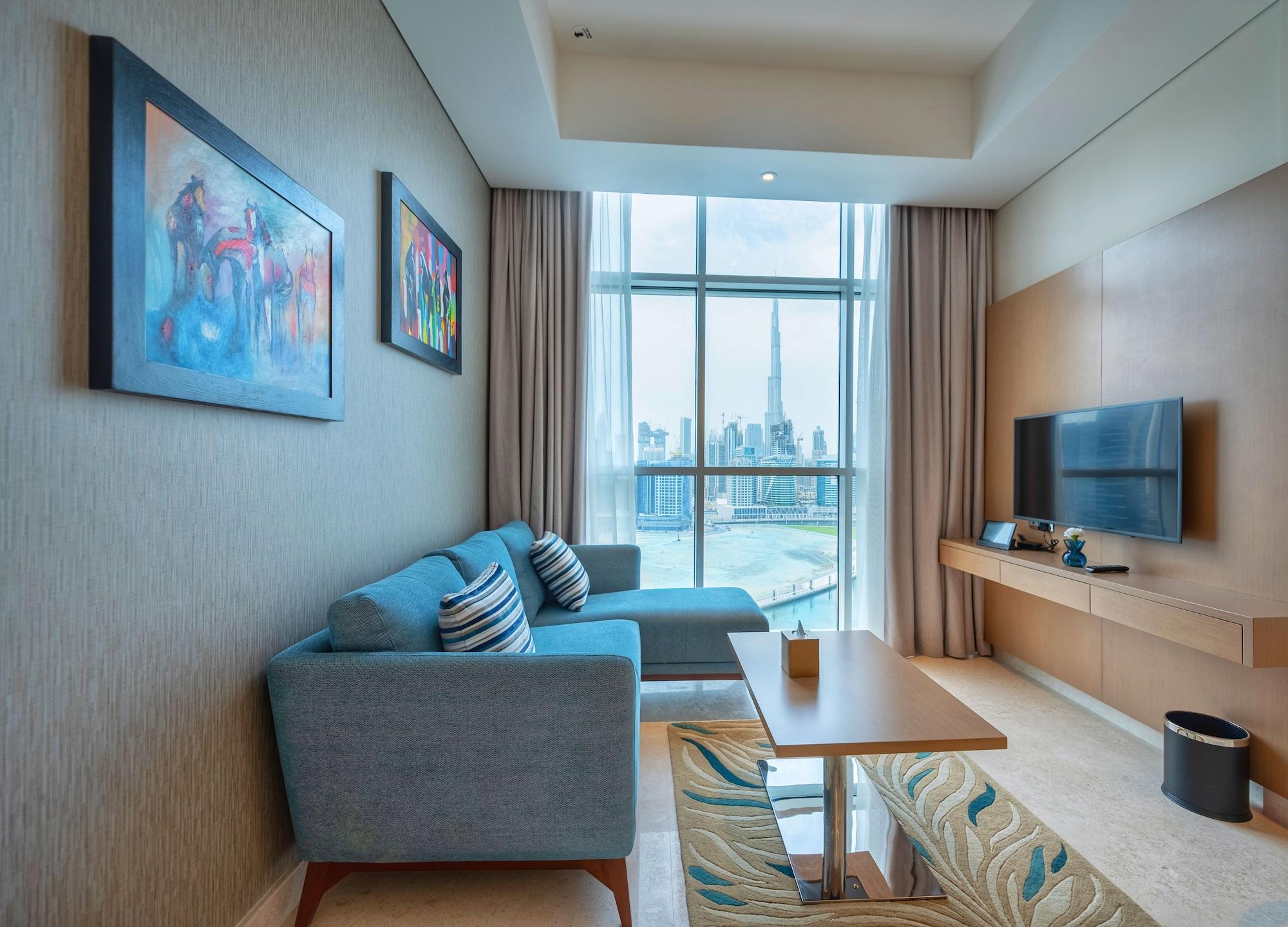 Canal Central Hotel Business Bay Dubaj Zewnętrze zdjęcie The photo shows a modern and stylish living room with large windows providing a view of a city skyline, including a notable tall building. The walls are adorned with colorful abstract paintings. There’s a light blue sofa with decorative cushions, and