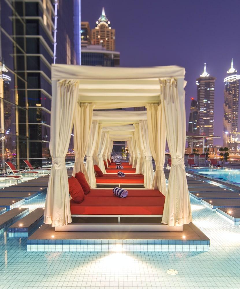Canal Central Hotel Business Bay Dubaj Zewnętrze zdjęcie The photo shows a luxurious outdoor lounge area featuring several cabanas with sheer white curtains. Each cabana has a red sofa with cushions, arranged in a symmetrical fashion along a pathway. The setting appears to be near a pool, illuminated by so