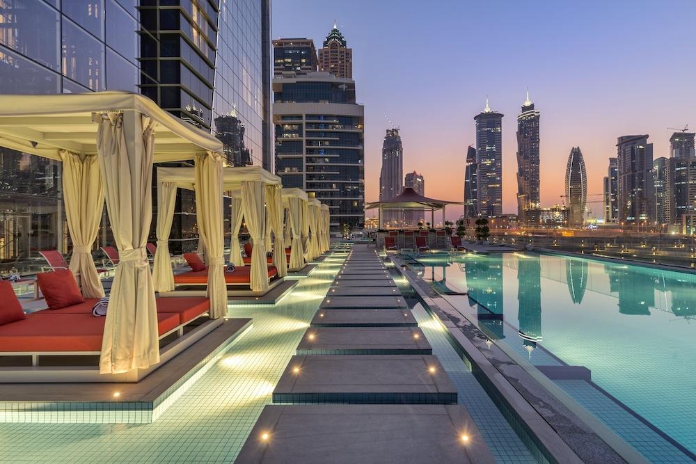 Canal Central Hotel Business Bay Dubaj Zewnętrze zdjęcie The photo shows a modern, luxurious outdoor space featuring a long swimming pool lined with sleek lounge chairs and shaded cabanas. The cabanas are draped with light-colored fabric and have vibrant red cushions. On either side of the pool, there are 