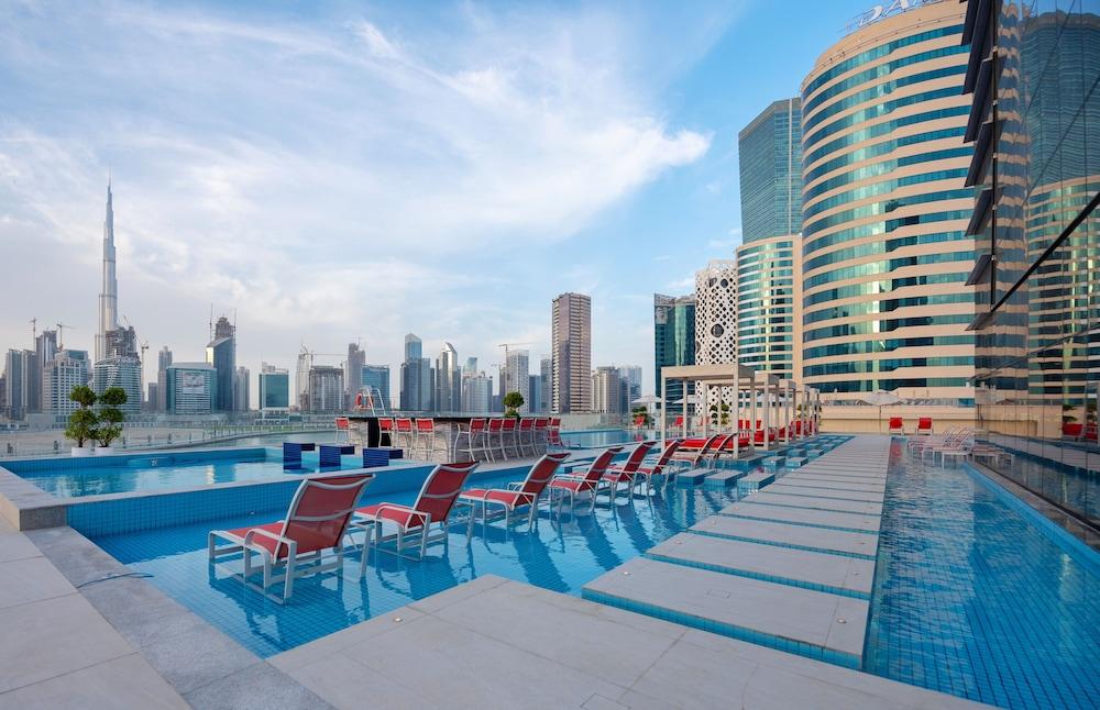 Canal Central Hotel Business Bay Dubaj Zewnętrze zdjęcie The photo depicts a modern swimming pool surrounded by skyscrapers in an urban setting. The pool features a sleek design with lounge chairs arranged along its edge. In the background, various tall buildings, including one that appears to be the Burj 