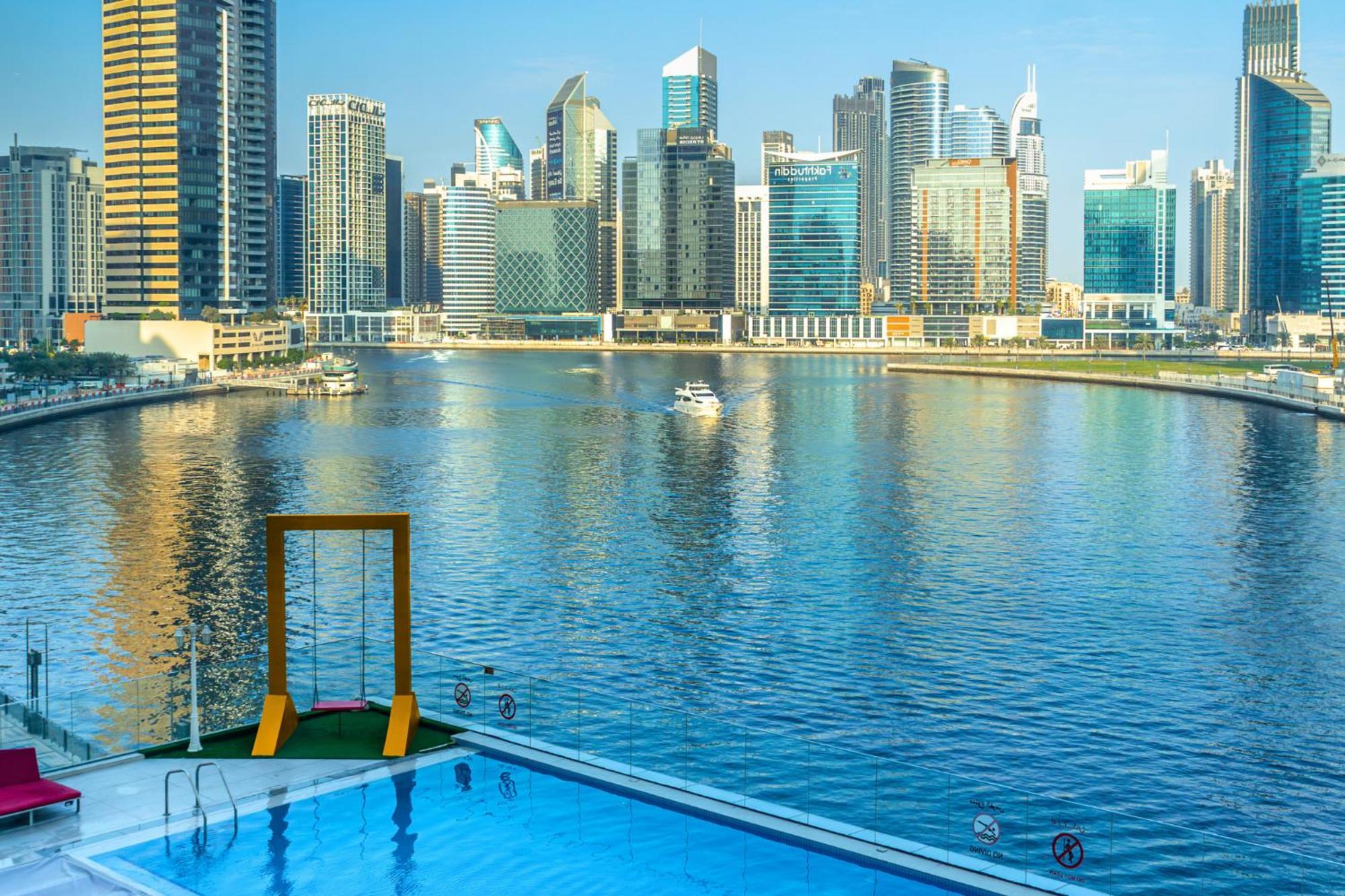 Canal Central Hotel Business Bay Dubaj Zewnętrze zdjęcie The photo shows a serene waterfront view featuring a modern city skyline with tall buildings reflecting in the water. In the foreground, there is a swimming pool with a swing set beside it, overlooking the calm water. The skyline is characterized by 