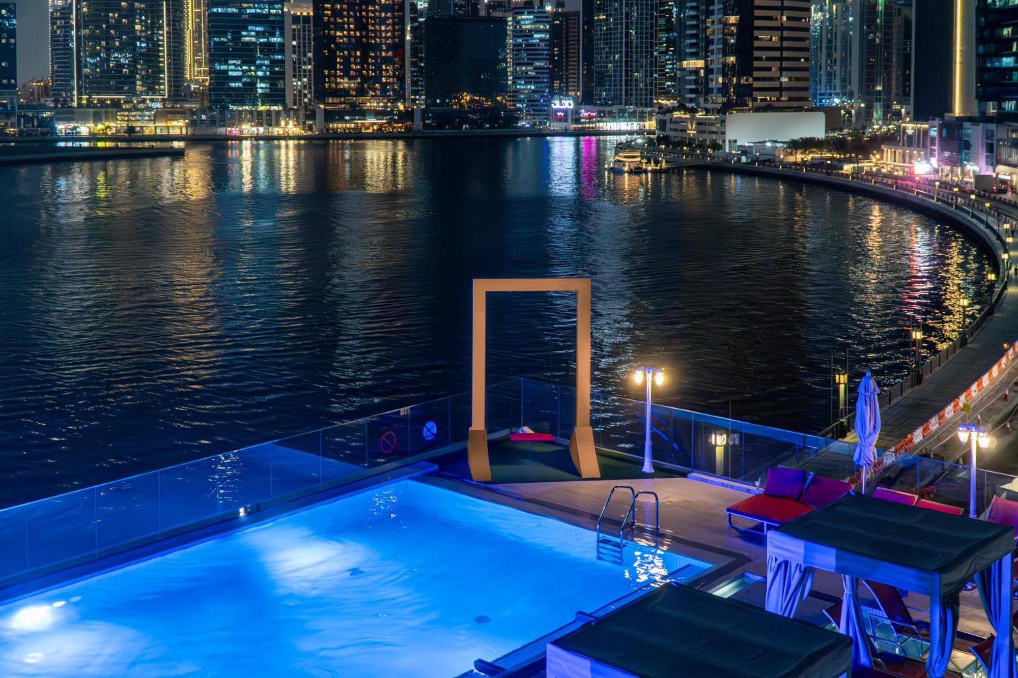 Canal Central Hotel Business Bay Dubaj Zewnętrze zdjęcie The photo shows a nighttime view of a modern pool area overlooking a body of water, likely a marina or waterfront. The pool is illuminated with a blue light, and there are lounge chairs beside it. In the background, a city skyline can be seen with ta