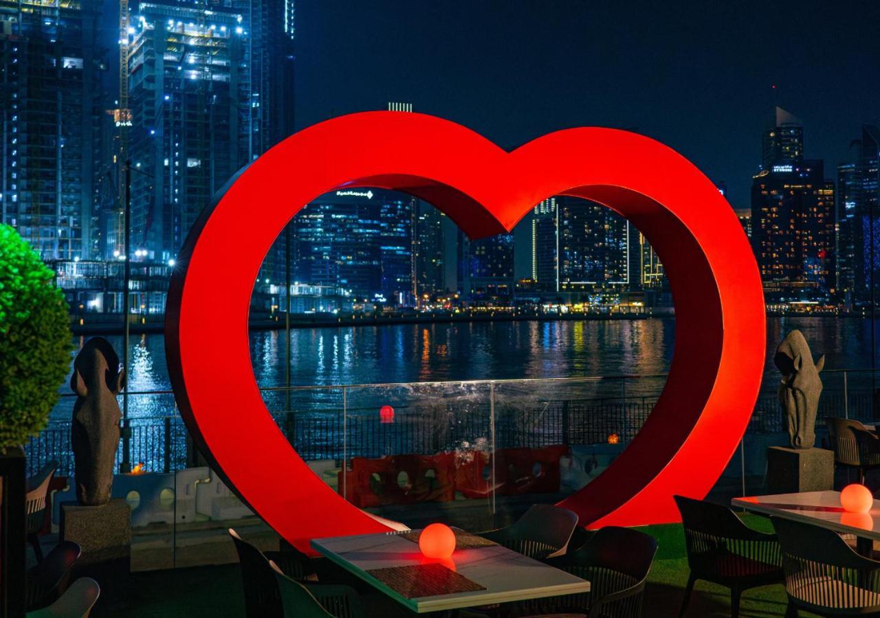 Canal Central Hotel Business Bay Dubaj Zewnętrze zdjęcie The photo depicts a large, illuminated red heart sculpture set against a nighttime urban backdrop. The heart is prominently displayed near a body of water, with city skyline buildings and lights reflecting off the surface. There are tables in the for