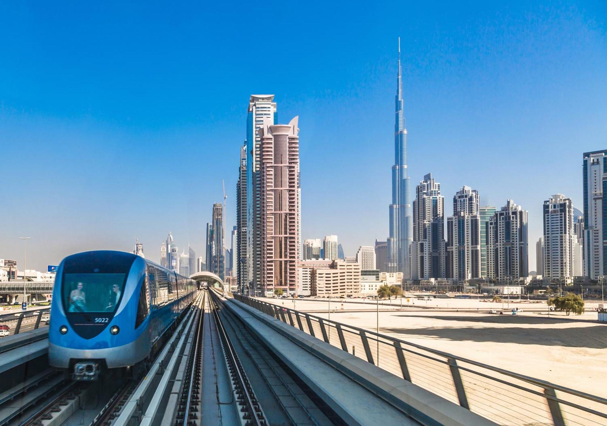 Canal Central Hotel Business Bay Dubaj Zewnętrze zdjęcie The photo shows a modern metro train on elevated tracks, traveling through a cityscape. To the right, there's a cluster of tall skyscrapers, one of which is recognizable as the Burj Khalifa, the tallest building in the world. The sky is clear and blu