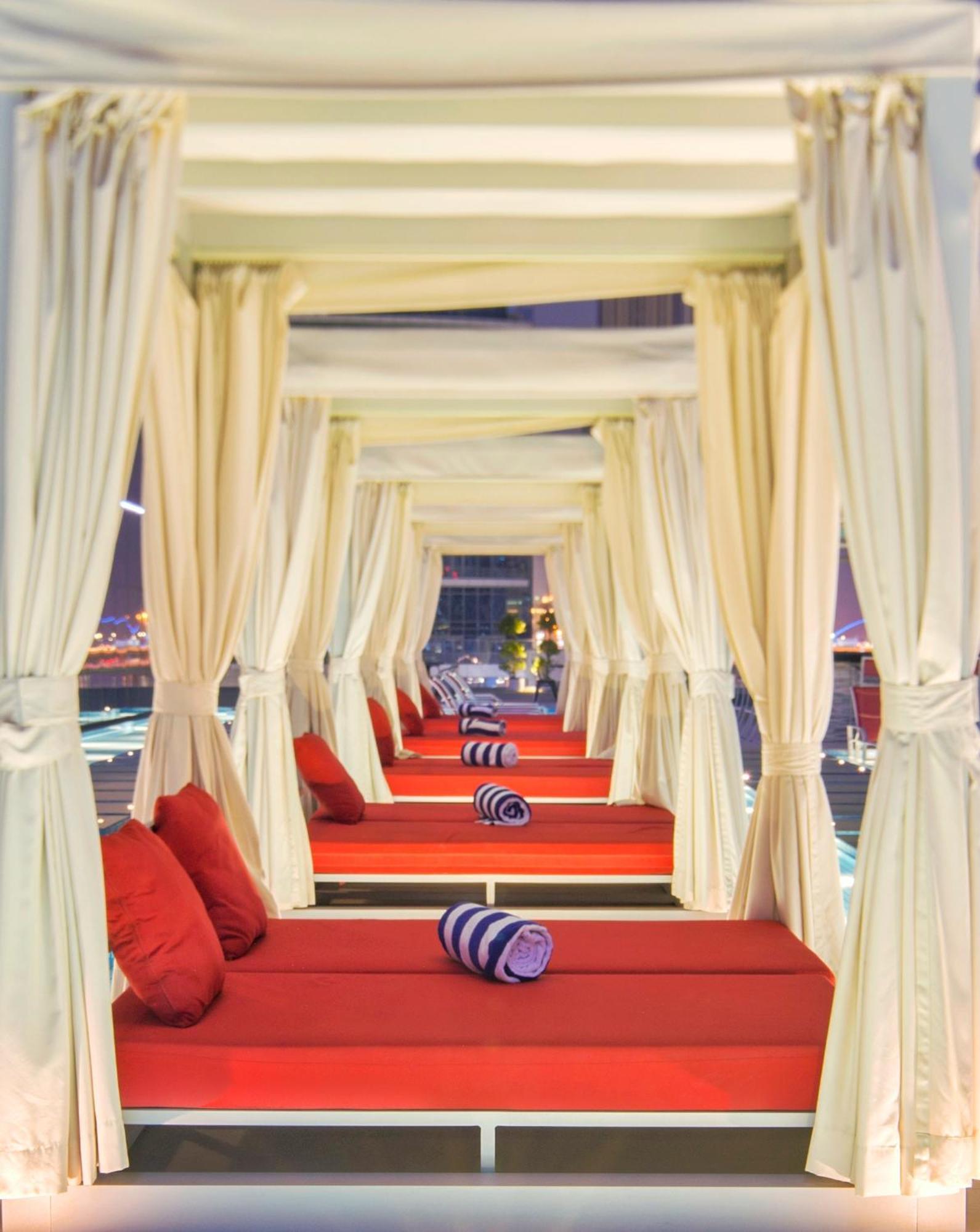Canal Central Hotel Business Bay Dubaj Zewnętrze zdjęcie The photo shows a series of elegant lounging areas, likely in a luxurious outdoor setting. Each area is framed by white curtains, creating a cozy, intimate atmosphere. The lounging beds are cushioned with red fabric and feature rolled towels for rela
