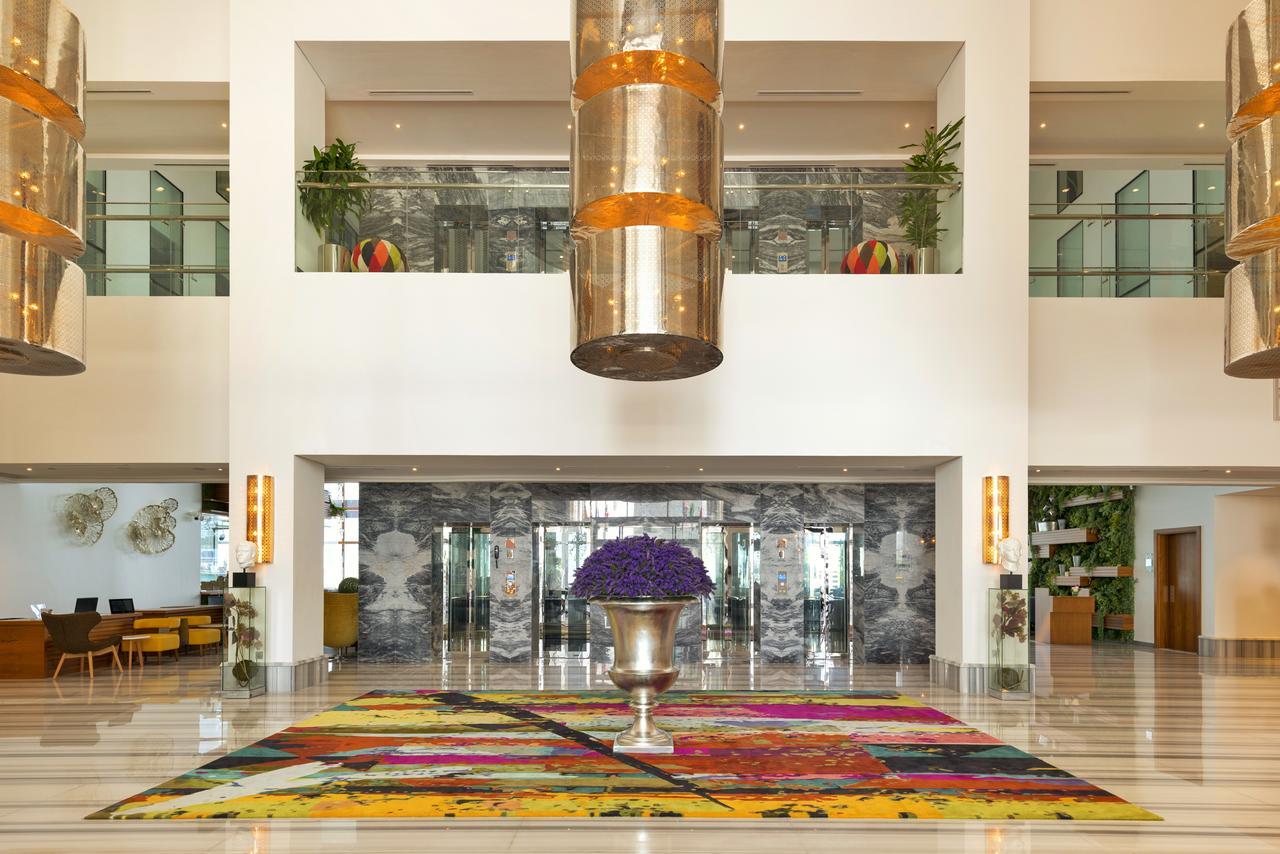 Canal Central Hotel Business Bay Dubaj Zewnętrze zdjęcie The photo shows a spacious and modern hotel lobby. Central to the scene is a large, vibrant area rug featuring a colorful abstract design with various hues. In the middle of the rug, there is a decorative metallic planter filled with purple flowers. 