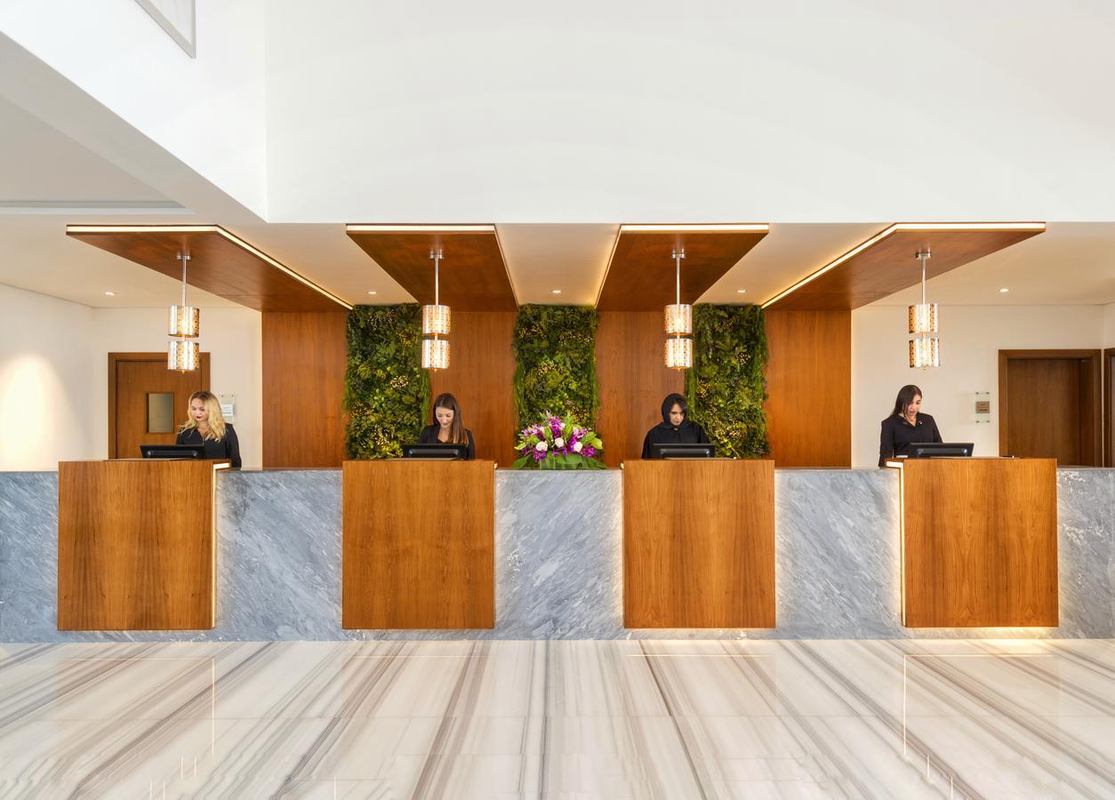 Canal Central Hotel Business Bay Dubaj Zewnętrze zdjęcie The photo shows a hotel reception area with a modern design. There are four reception desks, each made of wood and marble. Behind the desks, there are green plants as part of the décor, adding a fresh touch. The staff at the desks appear to be engagi