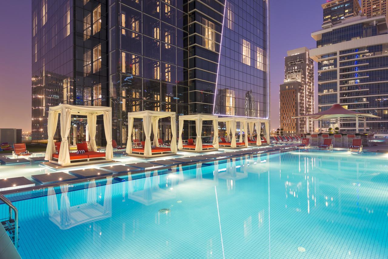 Canal Central Hotel Business Bay Dubaj Zewnętrze zdjęcie The photo shows a modern outdoor pool area at dusk, with a clear blue pool reflecting the surrounding buildings. White cabanas with red cushions line the edge of the pool, providing a comfortable lounging space. The background features tall, sleek sk