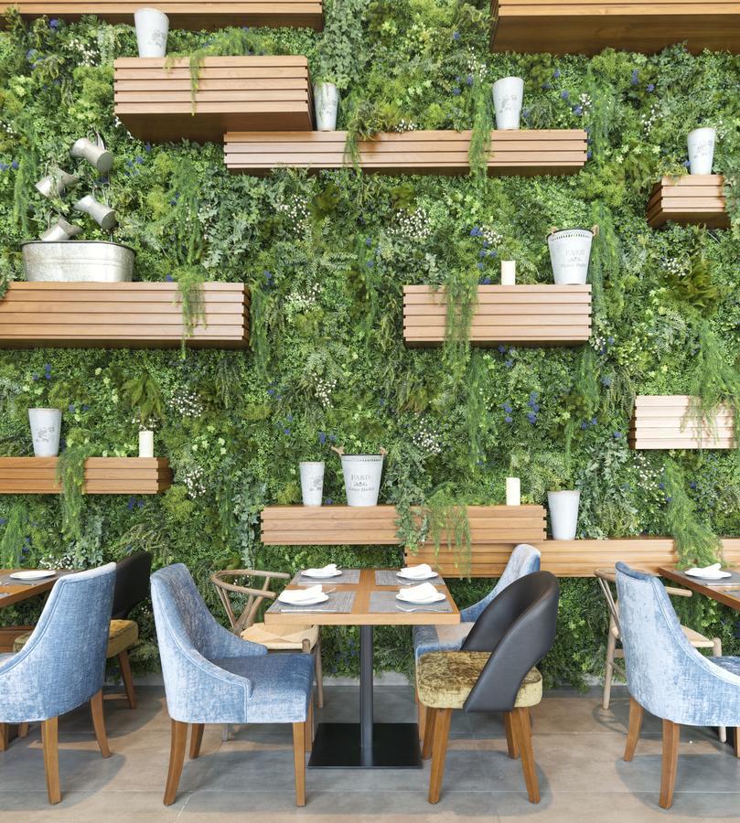Canal Central Hotel Business Bay Dubaj Zewnętrze zdjęcie The photo depicts a vibrant interior of a restaurant featuring a green wall adorned with various plants. There are wooden shelves mounted on the wall, displaying decorative white pots and planters. The setting includes several tables with neatly arra