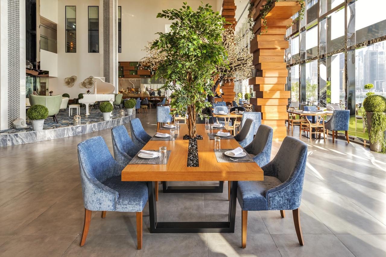 Canal Central Hotel Business Bay Dubaj Zewnętrze zdjęcie The photo shows a modern restaurant interior featuring a spacious dining area. In the center, there is a large wooden table surrounded by upholstered blue chairs. A tall, green plant is positioned in the middle of the table, adding a natural element 