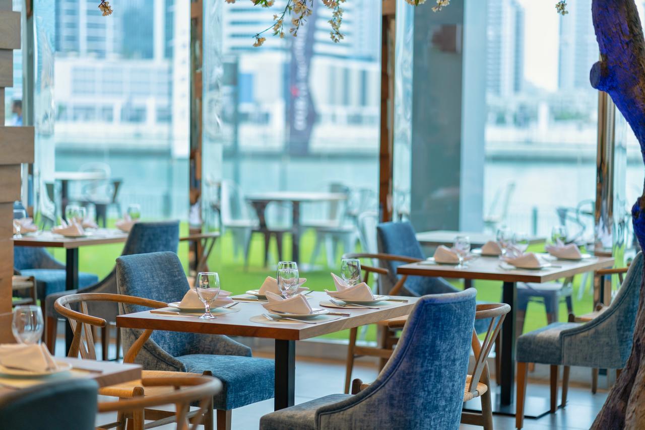 Canal Central Hotel Business Bay Dubaj Zewnętrze zdjęcie The image shows a modern restaurant interior featuring tables set with dinnerware, glasses, and napkins. The furnishings include elegant blue chairs and wooden tables. Large glass windows provide a view of the outdoors and a body of water, contributi