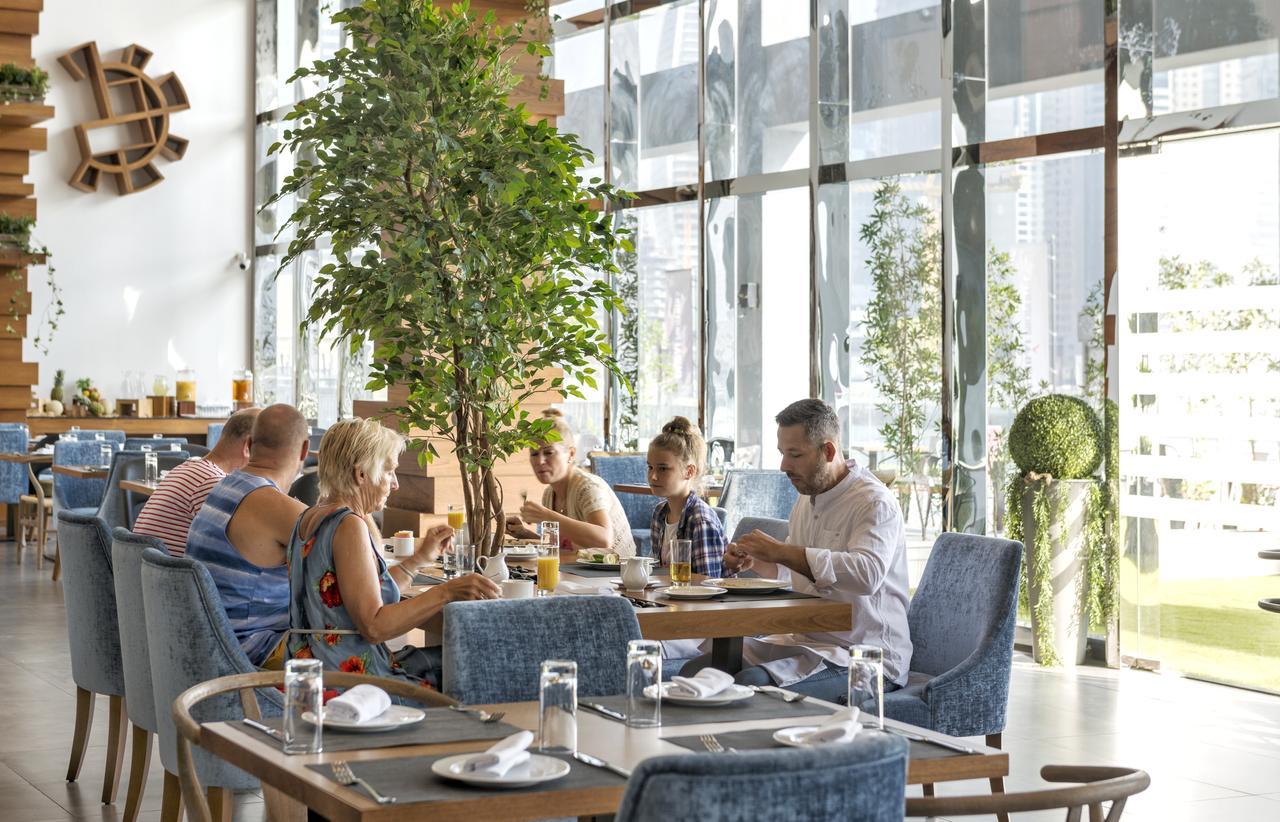 Canal Central Hotel Business Bay Dubaj Zewnętrze zdjęcie The photo depicts a modern restaurant setting with a group of people dining together. They are seated at a table with an appealing arrangement of light plates and glasses of water. The interior features large windows allowing natural light to flood t