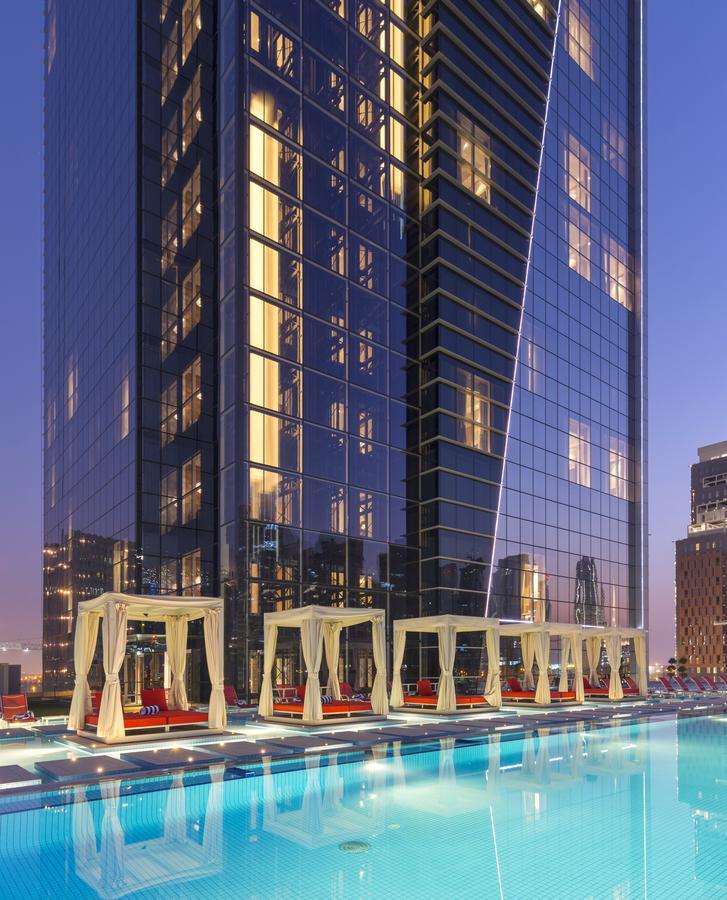 Canal Central Hotel Business Bay Dubaj Zewnętrze zdjęcie The image shows a modern skyscraper with a sleek glass facade, reflecting the surrounding cityscape. The building has prominent lighting, emphasizing its architectural design. In the foreground, there are several covered seating areas with white cano