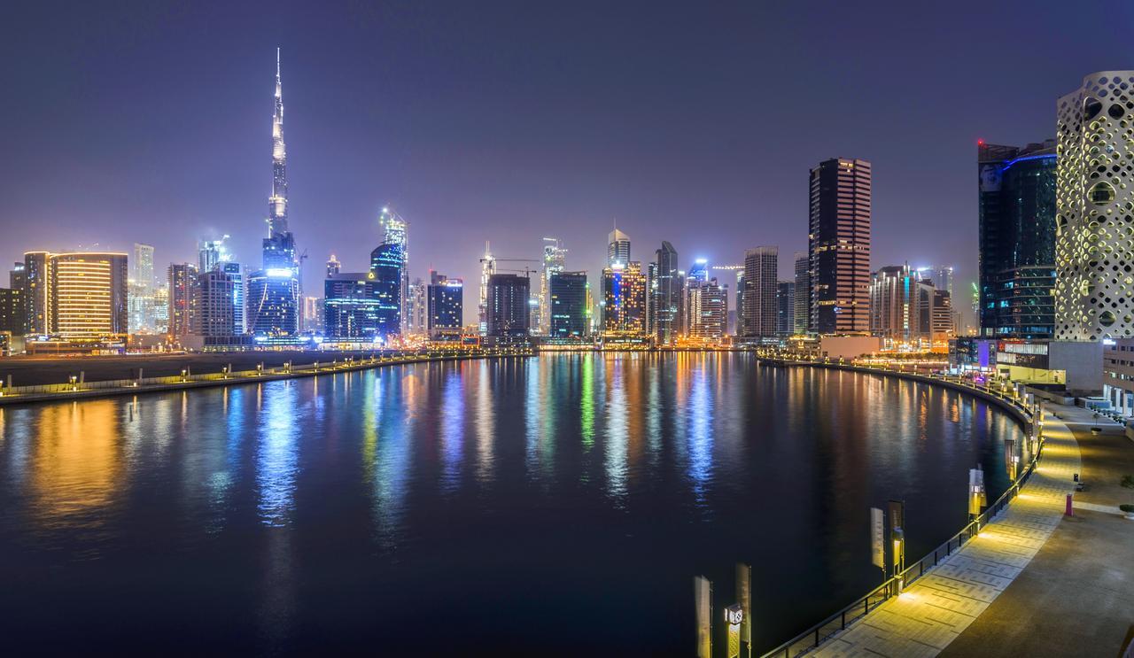 Canal Central Hotel Business Bay Dubaj Zewnętrze zdjęcie The photo shows a nighttime cityscape featuring a skyline illuminated by numerous lights. A prominent skyscraper, likely the Burj Khalifa, stands tall among other modern buildings. The scene includes a body of water reflecting the city lights, contri