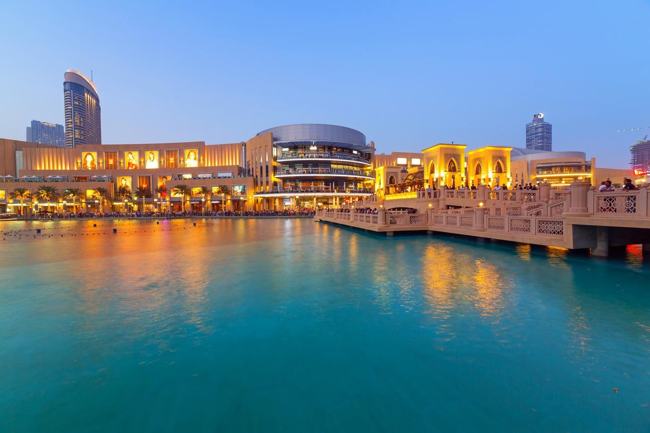 Canal Central Hotel Business Bay Dubaj Zewnętrze zdjęcie The photo depicts a scenic waterfront view, likely in an urban area during twilight. There's a large, modern building with bright lights, which seems to house shops or restaurants, alongside stylish architecture. The water in the foreground is calm a