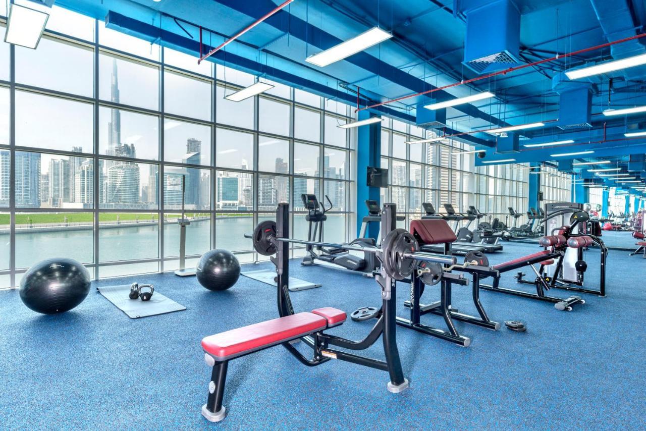 Canal Central Hotel Business Bay Dubaj Zewnętrze zdjęcie The photo shows a modern gym with a vibrant blue ceiling and walls. It features large windows that provide a view of a city skyline. Inside, there are various workout machines, including a bench press and cardio equipment. Additionally, there are exe